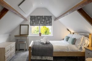 Cozy attic bedroom with wood beams, a comfortable bed, decorative throw, mirror, and desk by a window with floral blinds