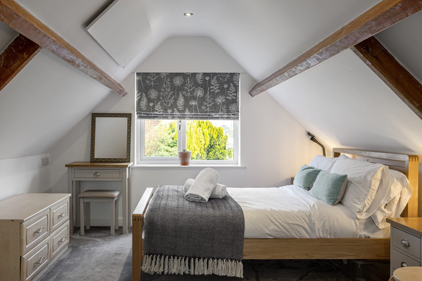 Cozy attic bedroom with wood beams, a comfortable bed, decorative throw, mirror, and desk by a window with floral blinds